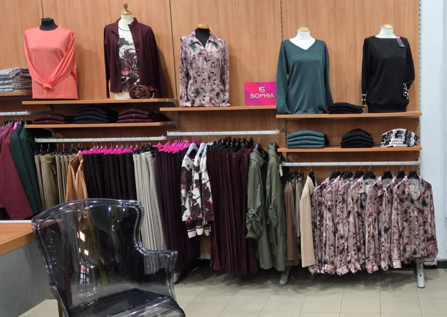 Angolo abbigliamento donna Sophia con maglie, camicie a fiori e pantaloni nei toni bordeaux, verde e rosa su manichini e stender